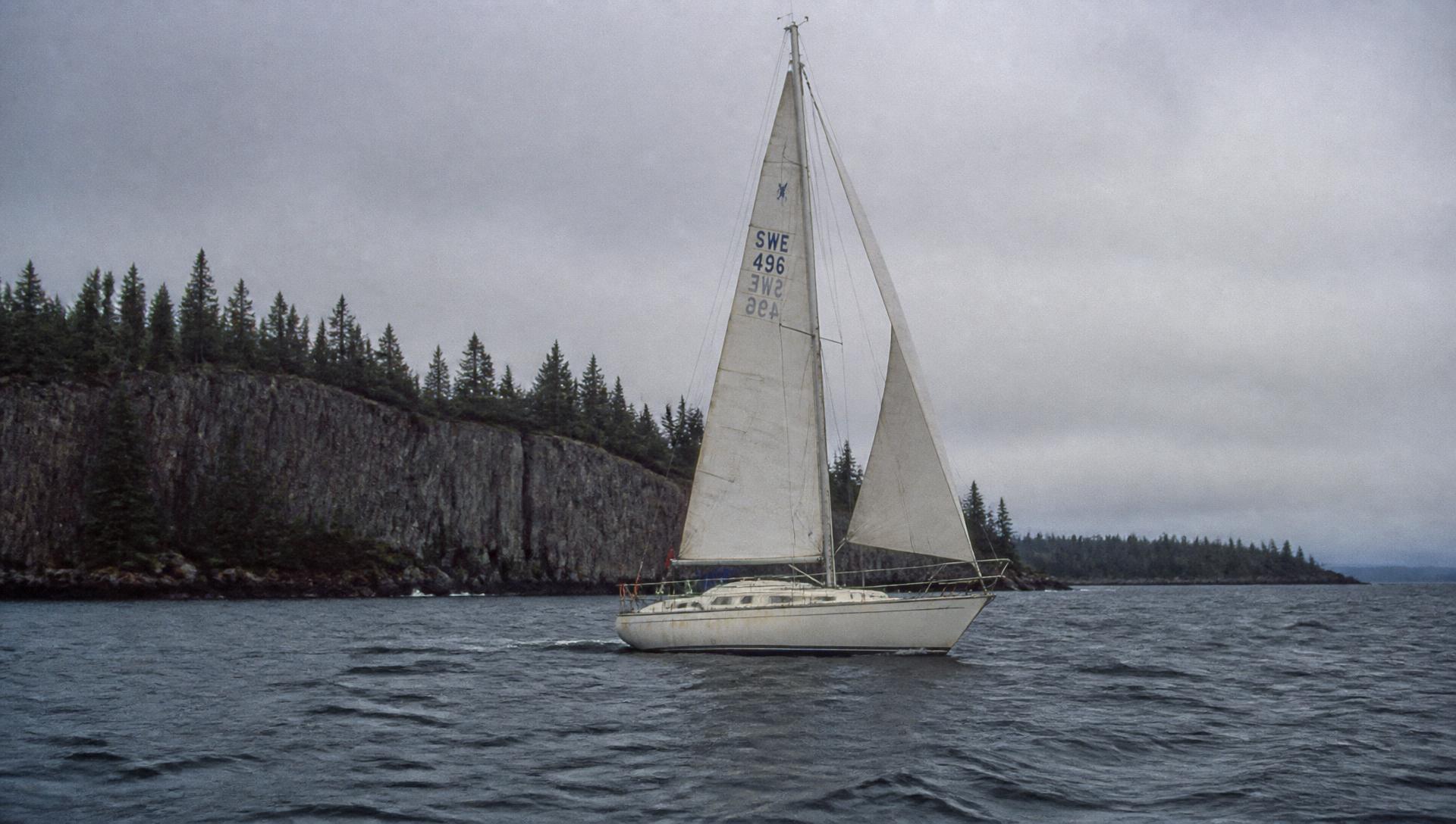 A small Swedish-built sloop sailing under overcast light along a rocky Lake Superior shoreline